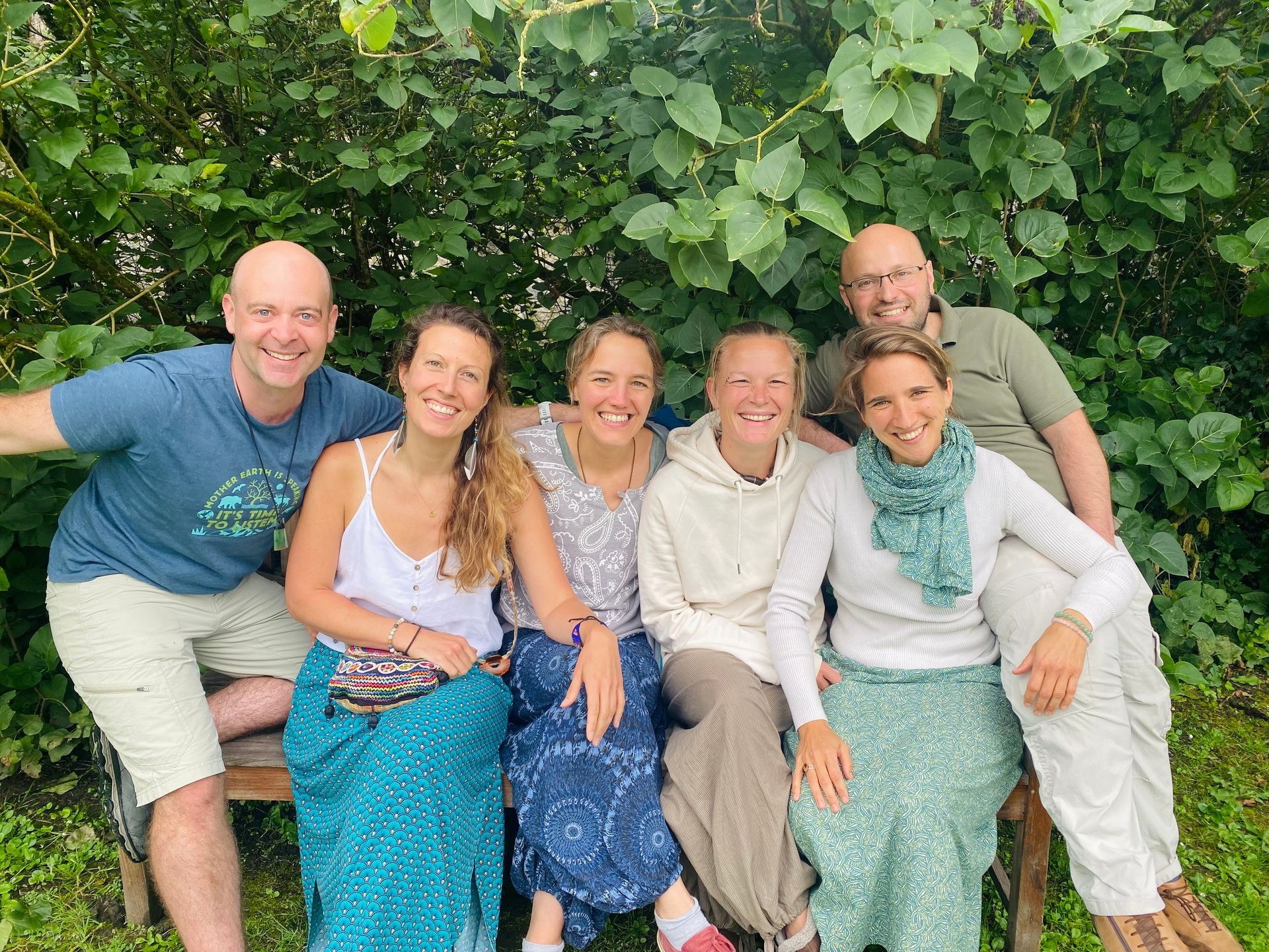 Group of six smiling people sitting on a bench in front of leafy green plants.