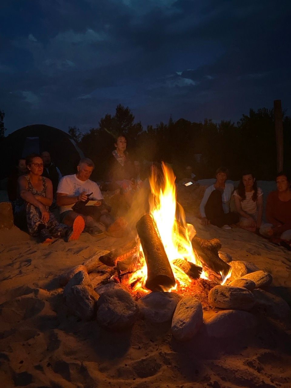 Group of people sitting around a campfire on a sandy beach under a cloudy night sky.