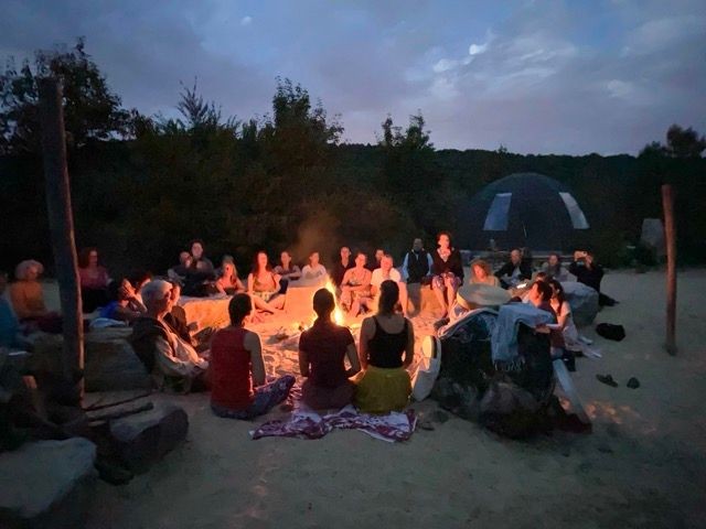 Group of people sitting around a bonfire at dusk, surrounded by trees.