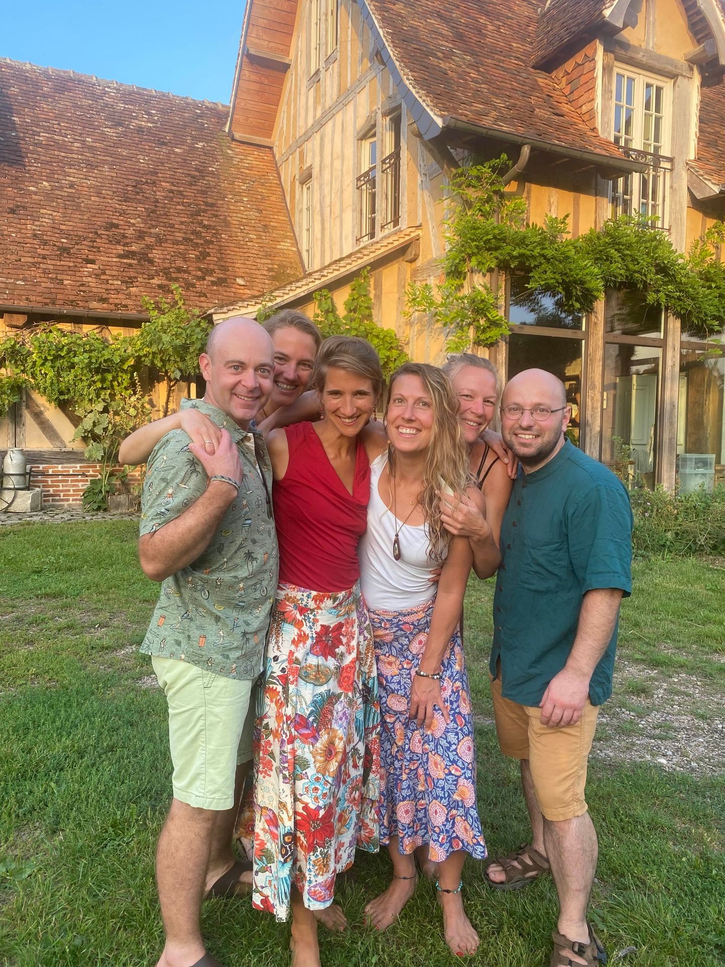 Group of people smiling and hugging in front of a rustic house with a sloped roof.